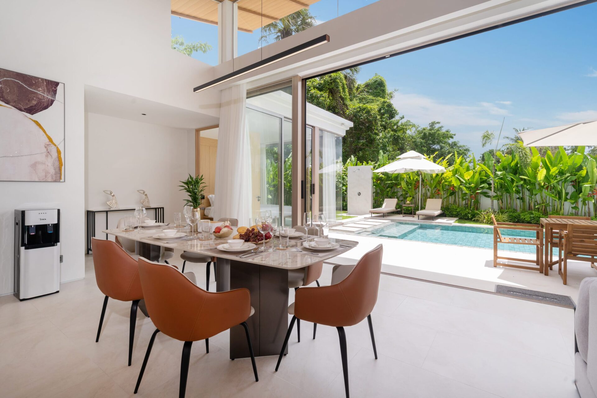 Open, sunlit dining area with a long table set for a meal, brown chairs, and glass sliding doors opening to a pool and tropical garden.