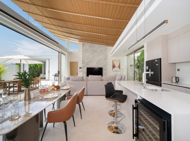 Open-plan kitchen and dining space with marble island, brown bar stools, and a glass wall leading to an outdoor patio with a white umbrella.