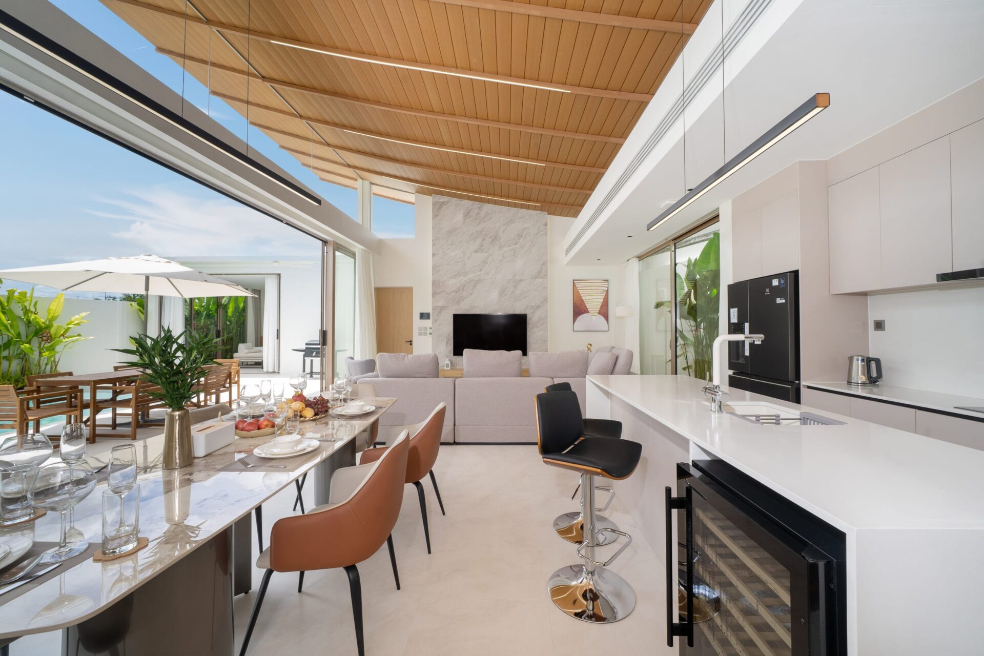 Open-plan kitchen and dining space with marble island, brown bar stools, and a glass wall leading to an outdoor patio with a white umbrella.