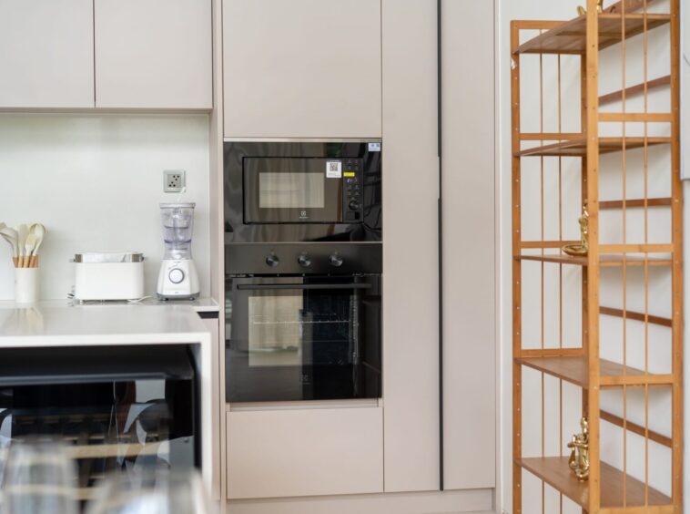 Beige kitchen with a built-in oven and microwave, white countertop, blender and toaster, and a wooden open shelving unit on the right.