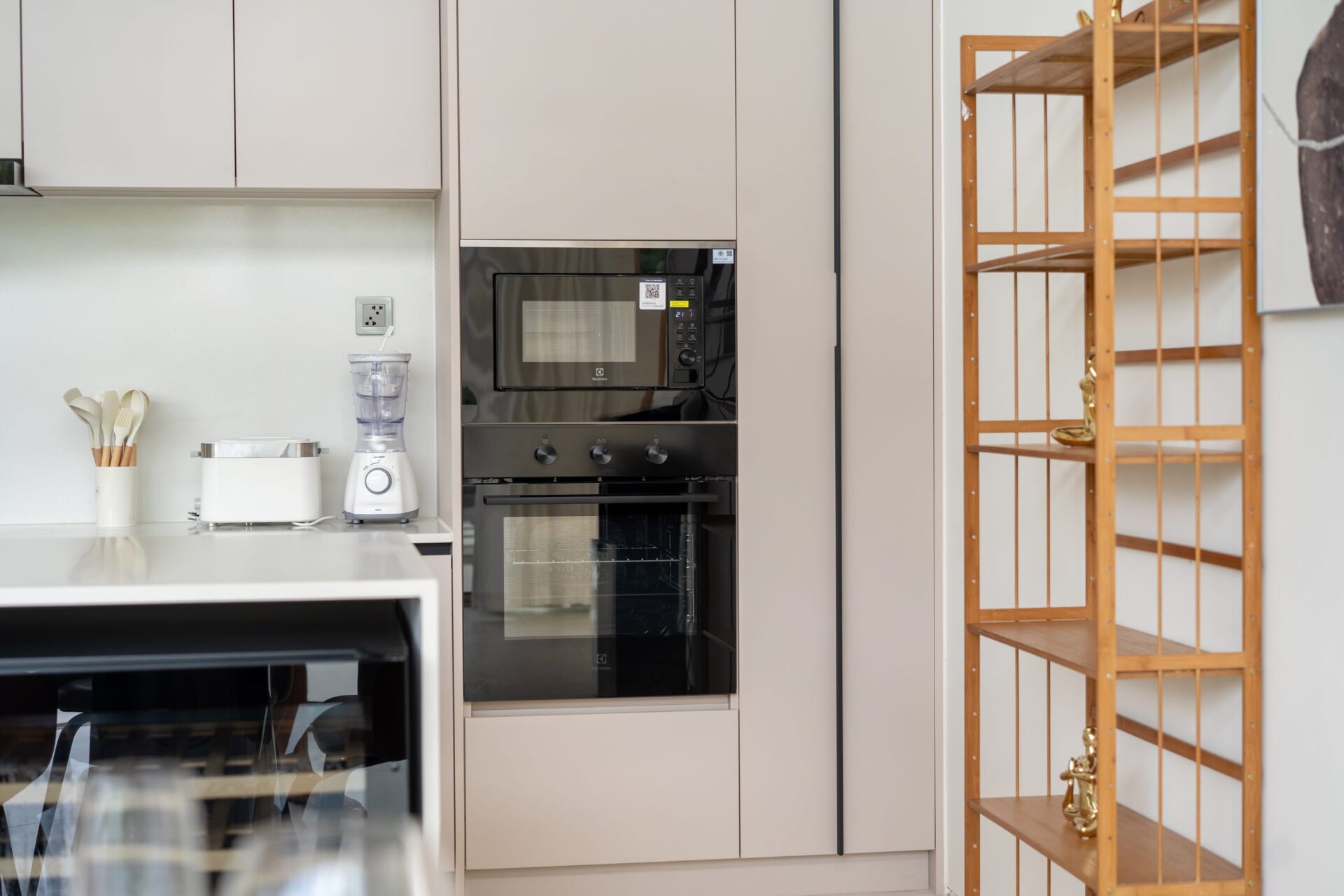 Beige kitchen with a built-in oven and microwave, white countertop, blender and toaster, and a wooden open shelving unit on the right.