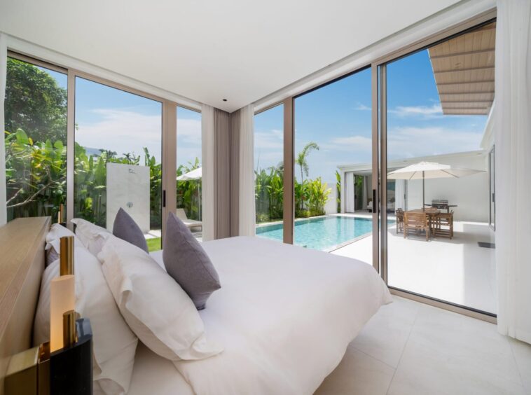 Luxurious hotel bedroom with floor-to-ceiling glass doors opening to a pool and tropical garden outside.