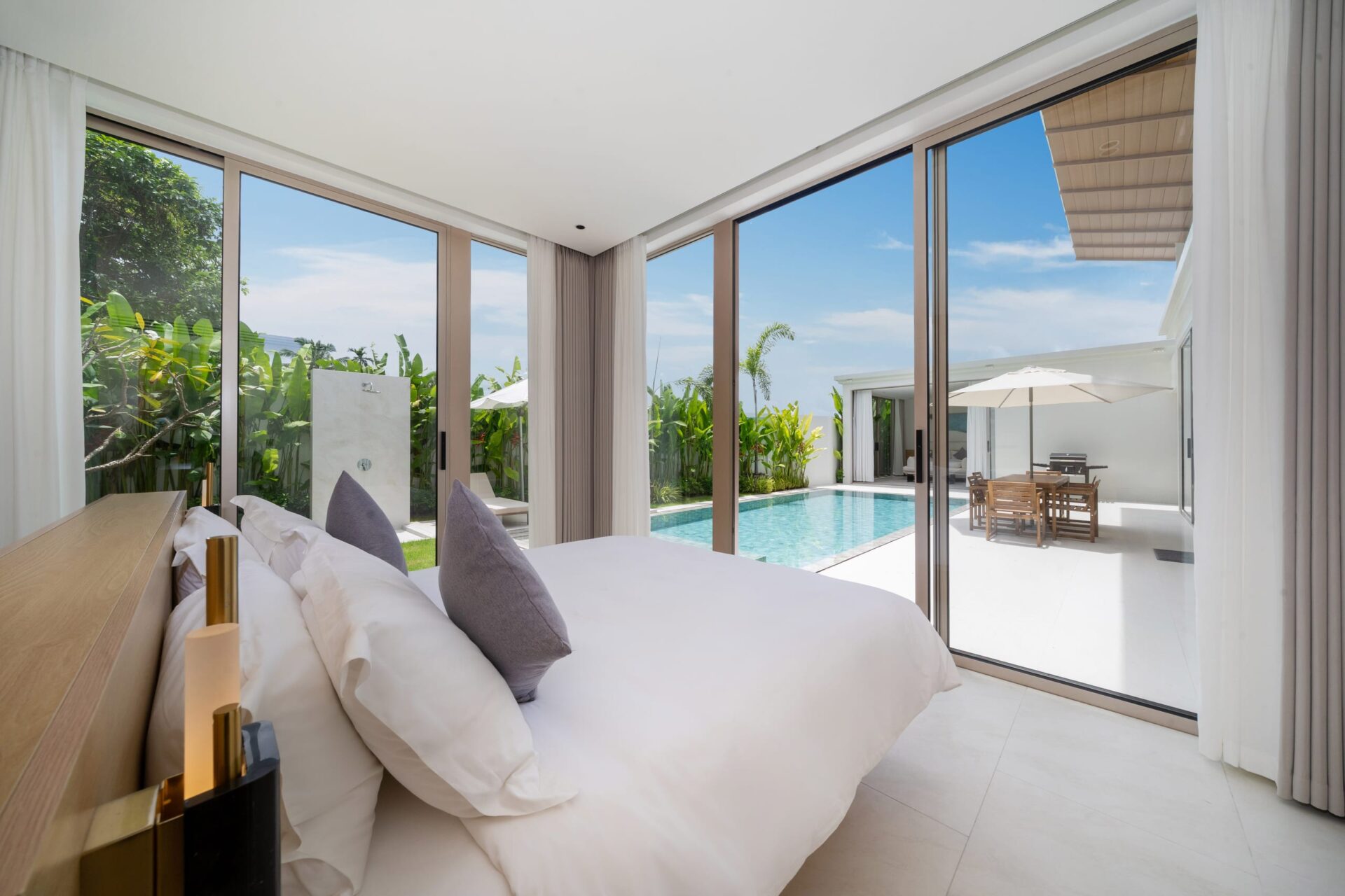 Luxurious hotel bedroom with floor-to-ceiling glass doors opening to a pool and tropical garden outside.