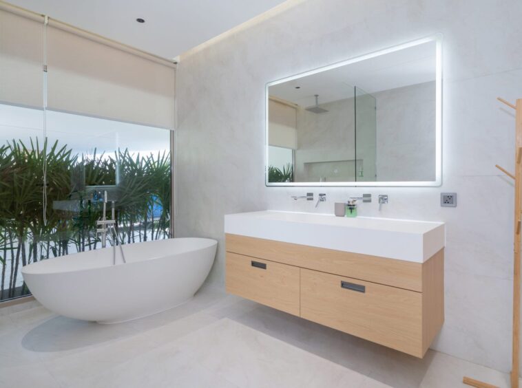 Modern bathroom with a freestanding oval tub beside a glass wall and plants, and a light wood vanity with a backlit mirror.