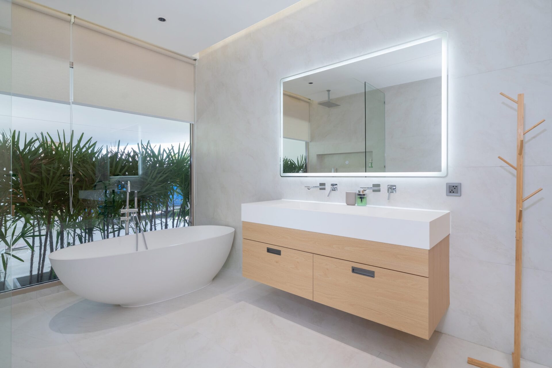 Modern bathroom with a freestanding oval tub beside a glass wall and plants, and a light wood vanity with a backlit mirror.