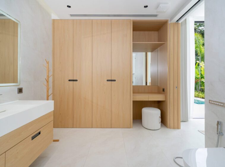 Spacious bathroom with light wood built-in wardrobes, white countertop sink, and a large framed mirror; a small round white stool sits near a built-in vanity, and sliding doors reveal an outdoor garden/pool area.