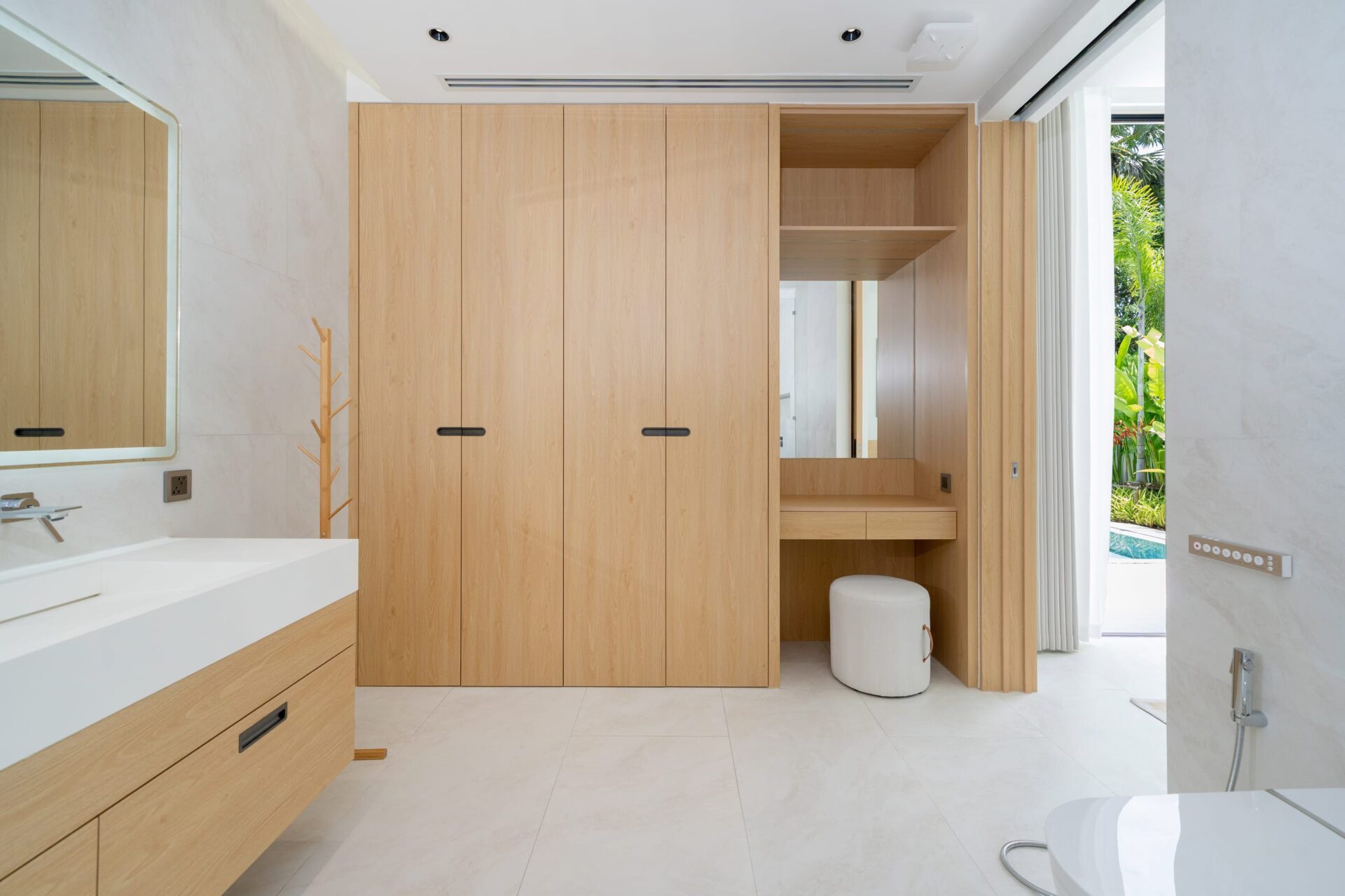 Spacious bathroom with light wood built-in wardrobes, white countertop sink, and a large framed mirror; a small round white stool sits near a built-in vanity, and sliding doors reveal an outdoor garden/pool area.