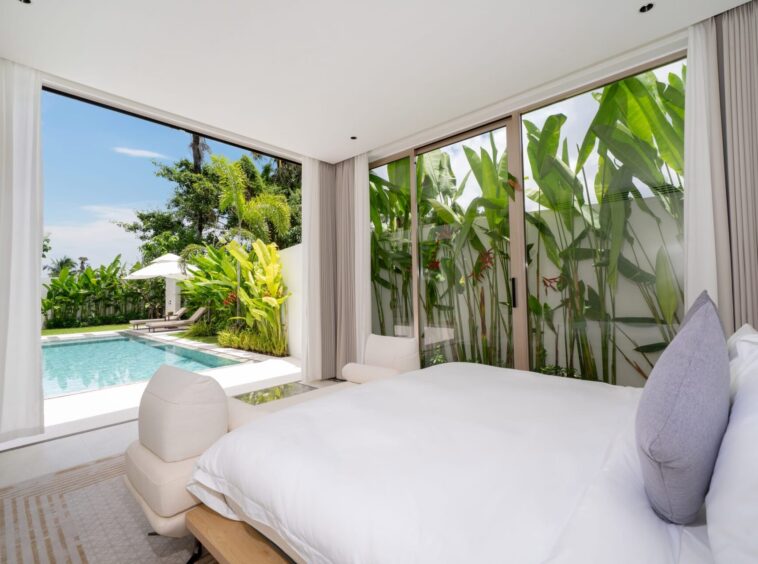 Bedroom with white bedding and gray pillows, open glass doors reveal a pool and tropical garden outside.