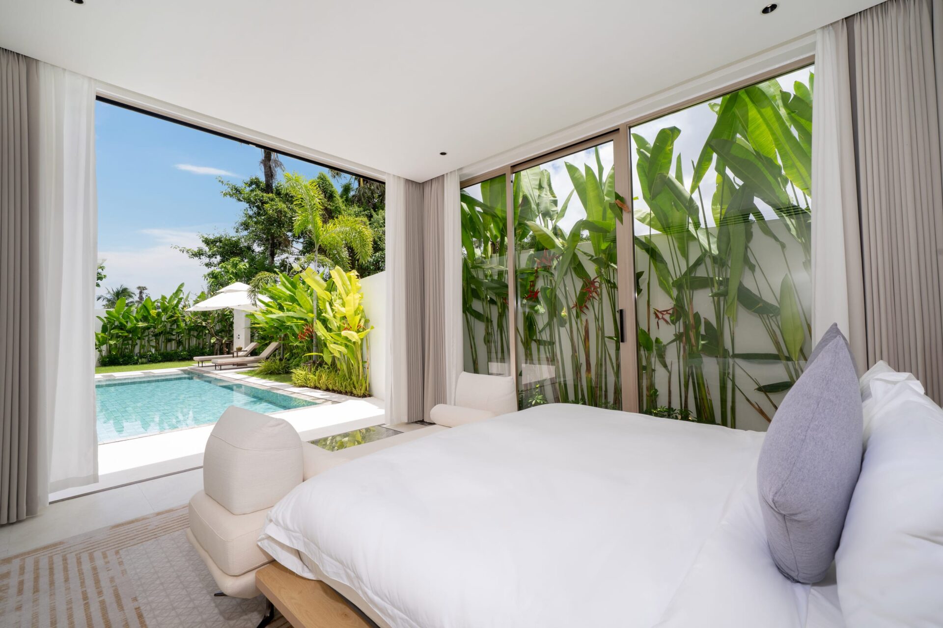 Bedroom with white bedding and gray pillows, open glass doors reveal a pool and tropical garden outside.