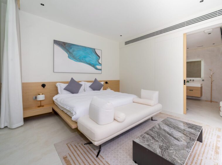 Modern hotel bedroom with a low wooden bed frame, white bedding and two gray accent pillows, a white chaise at the foot, and a marble-topped coffee table in front.