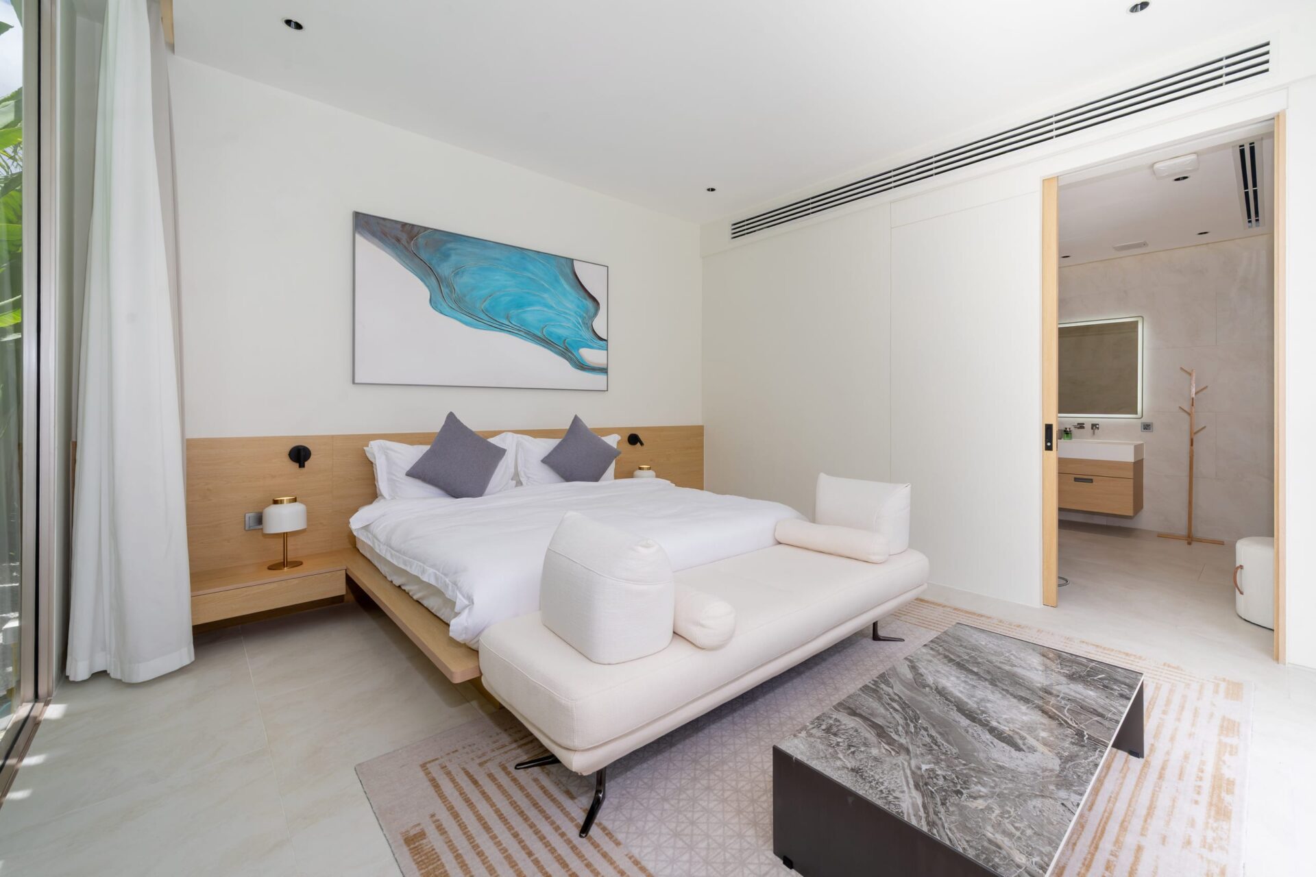 Modern hotel bedroom with a low wooden bed frame, white bedding and two gray accent pillows, a white chaise at the foot, and a marble-topped coffee table in front.