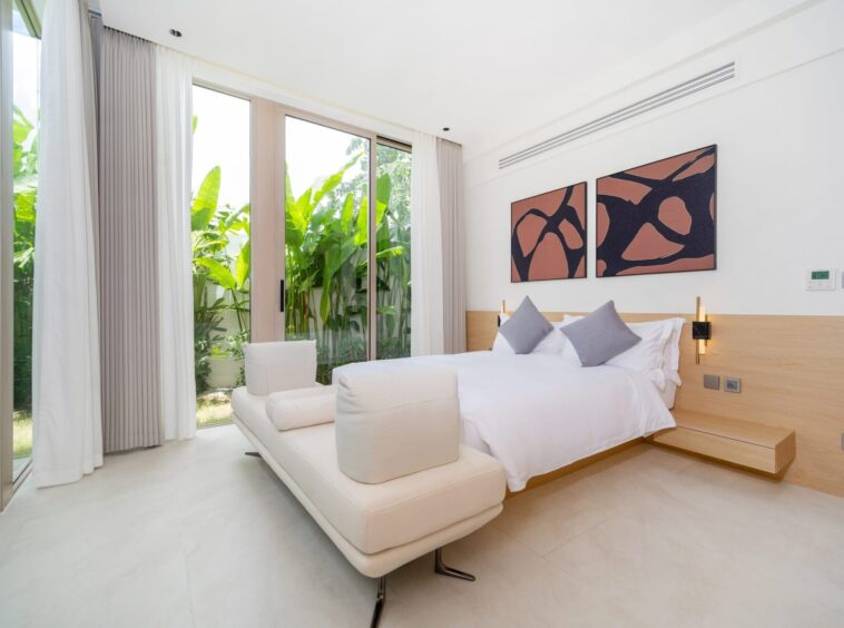 Modern bedroom with floor-to-ceiling glass doors opening to greenery, white bedding, and two abstract art panels above the bed.