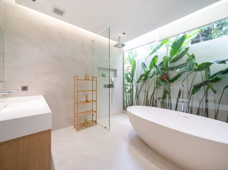 Bright, modern bathroom with a freestanding white tub, glass-enclosed shower, and a large glass wall overlooking tropical plants.