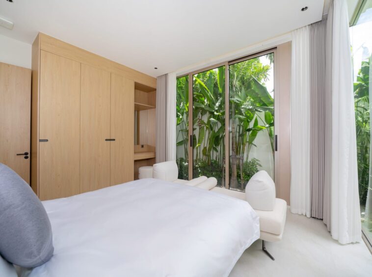 Minimalist bedroom with light wood wardrobes along the left wall and a glass sliding door to a tropical garden outside.