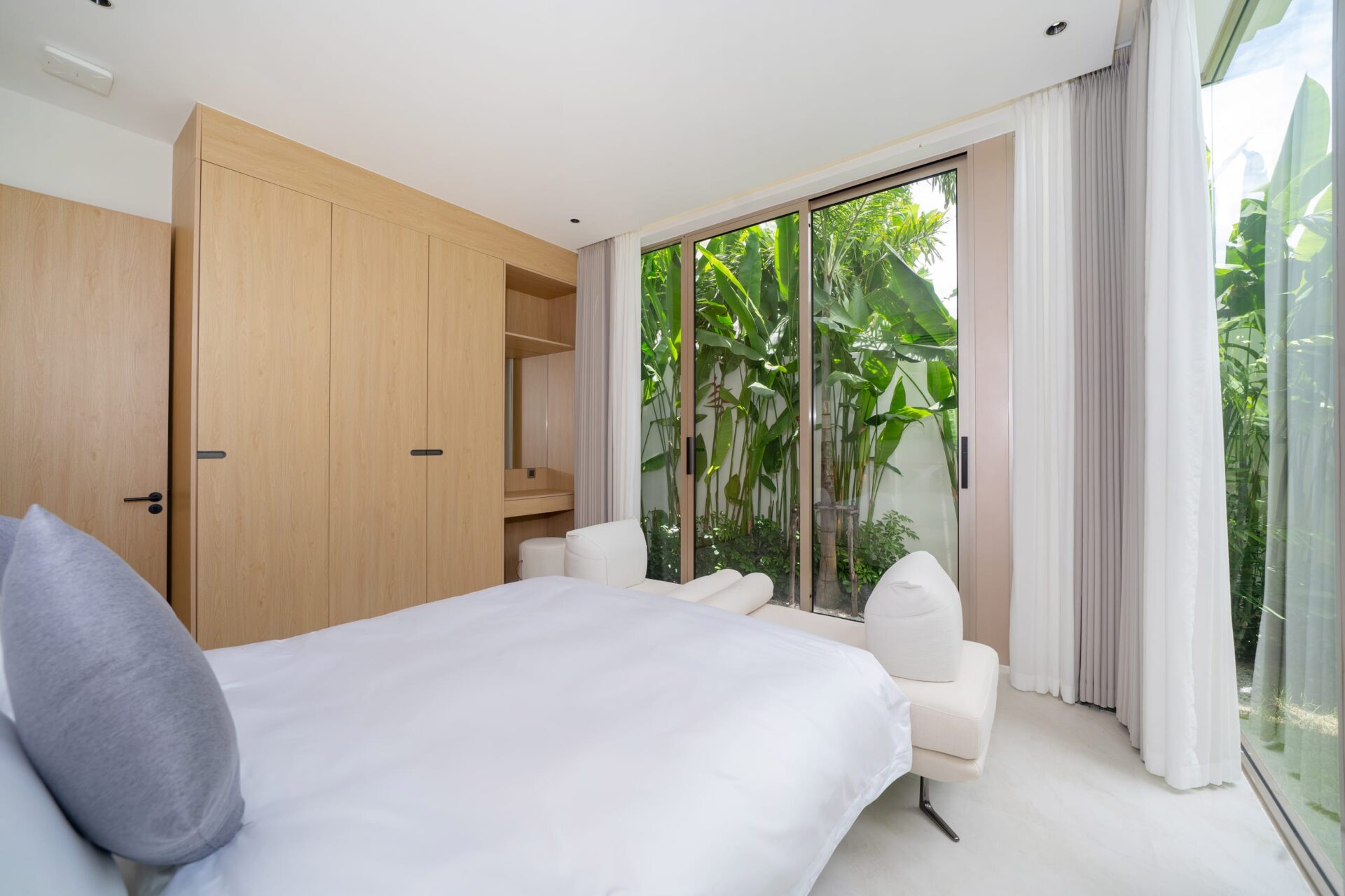 Minimalist bedroom with light wood wardrobes along the left wall and a glass sliding door to a tropical garden outside.