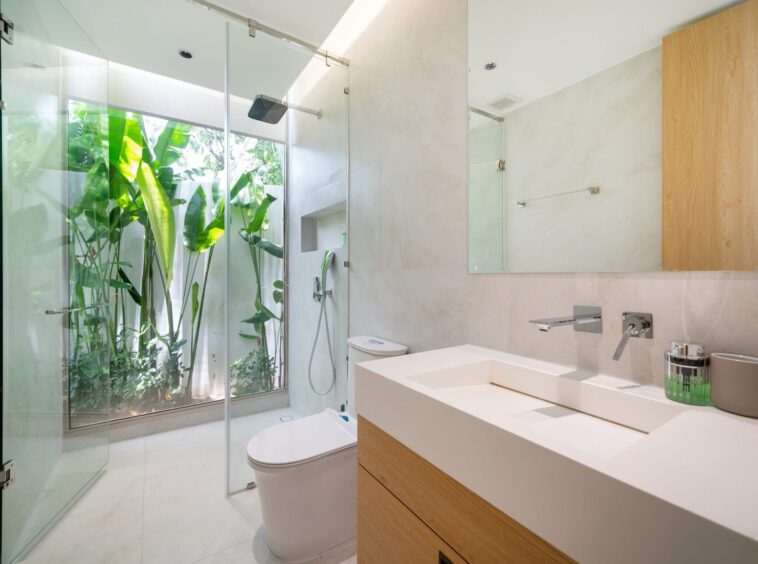 Contemporary bathroom with a glass shower enclosure, white double vanity, and lush green plants visible through the glass wall.