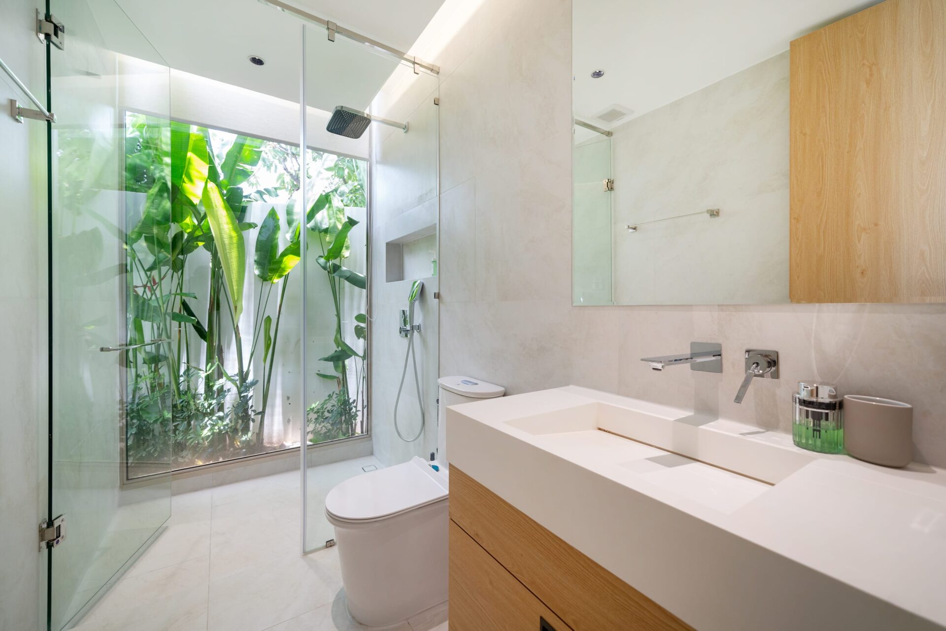 Contemporary bathroom with a glass shower enclosure, white double vanity, and lush green plants visible through the glass wall.