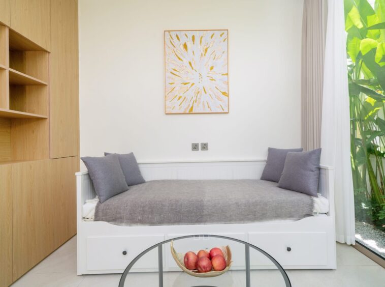 White daybed with gray cushions against a pale wall, with built-in wooden shelves on the left and a bright window with greenery on the right.
