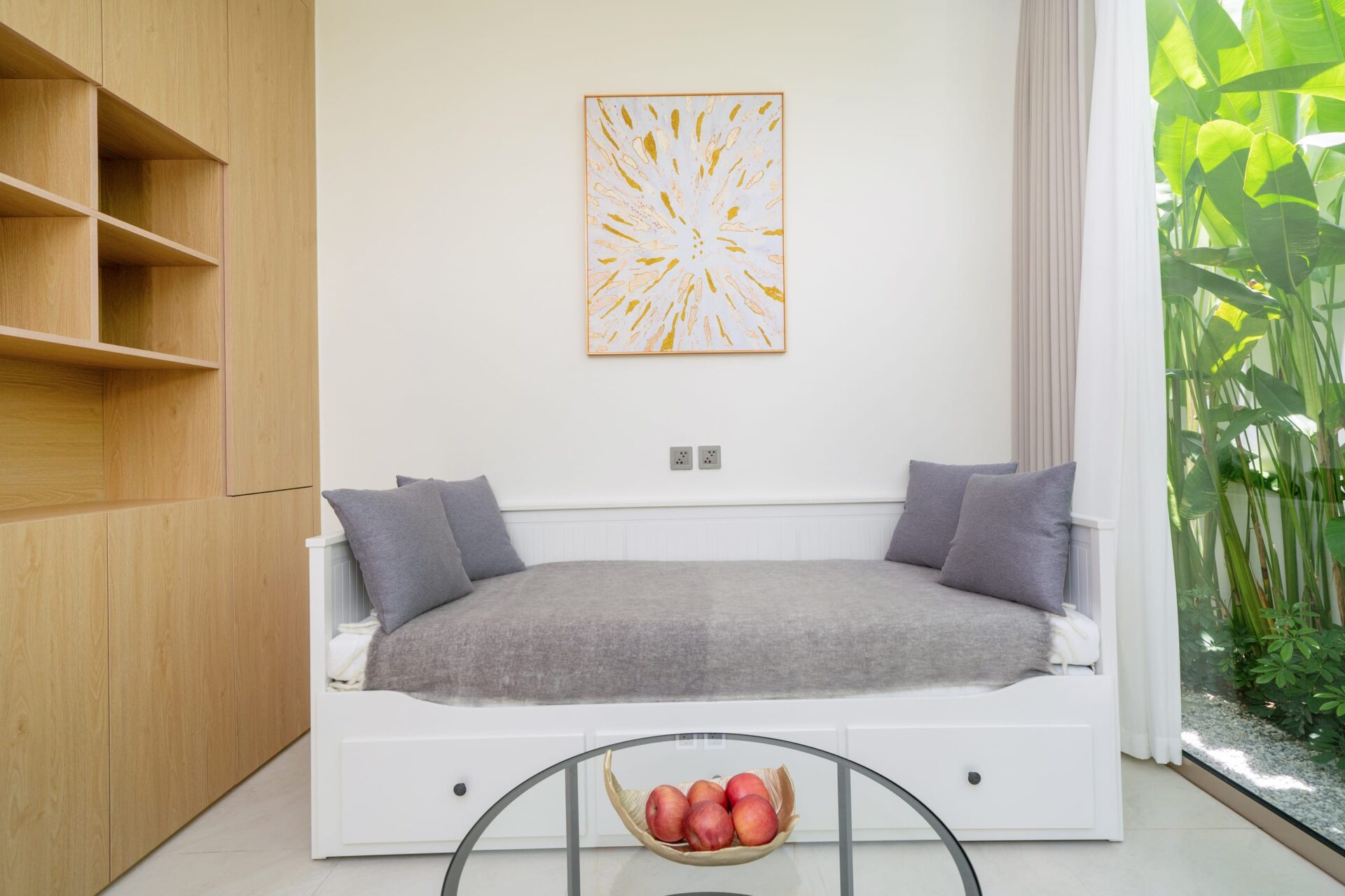 White daybed with gray cushions against a pale wall, with built-in wooden shelves on the left and a bright window with greenery on the right.