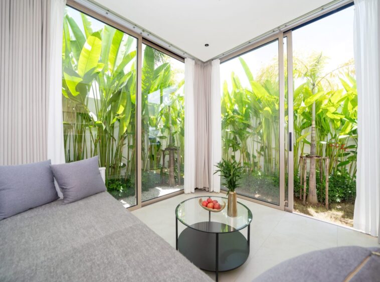 Bright sunroom with large glass sliding doors and lush tropical plants outside; gray sofa and round glass coffee table with fruit inside.