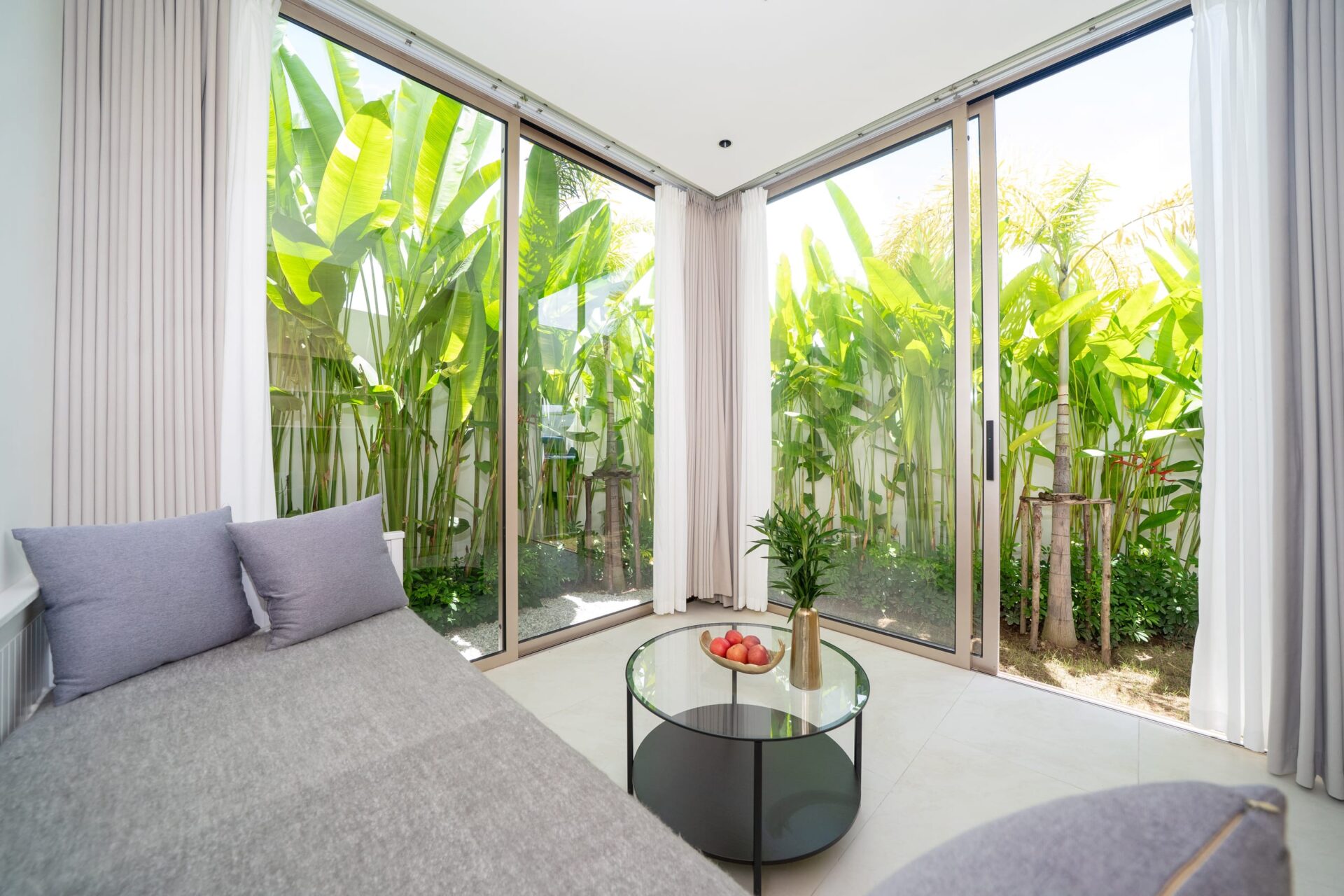 Bright sunroom with large glass sliding doors and lush tropical plants outside; gray sofa and round glass coffee table with fruit inside.