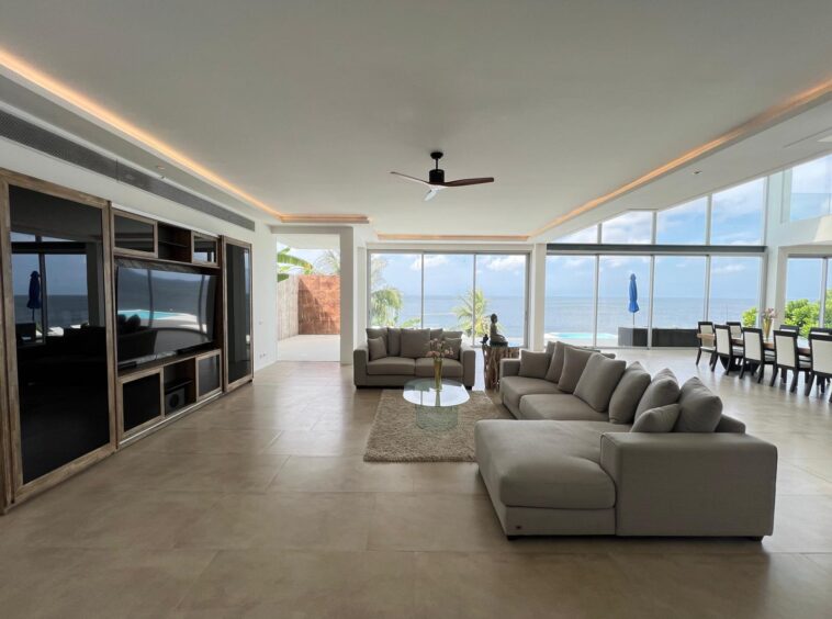 Open-plan living room with beige sofas facing a wall of windows overlooking the ocean; modern entertainment center on left and dining area to the right.