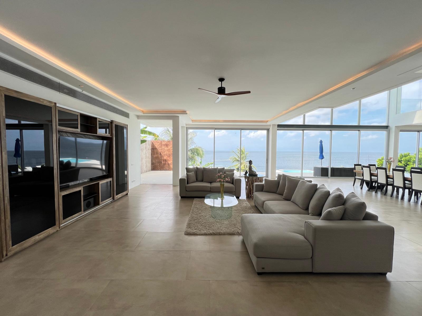 Open-plan living room with beige sofas facing a wall of windows overlooking the ocean; modern entertainment center on left and dining area to the right.