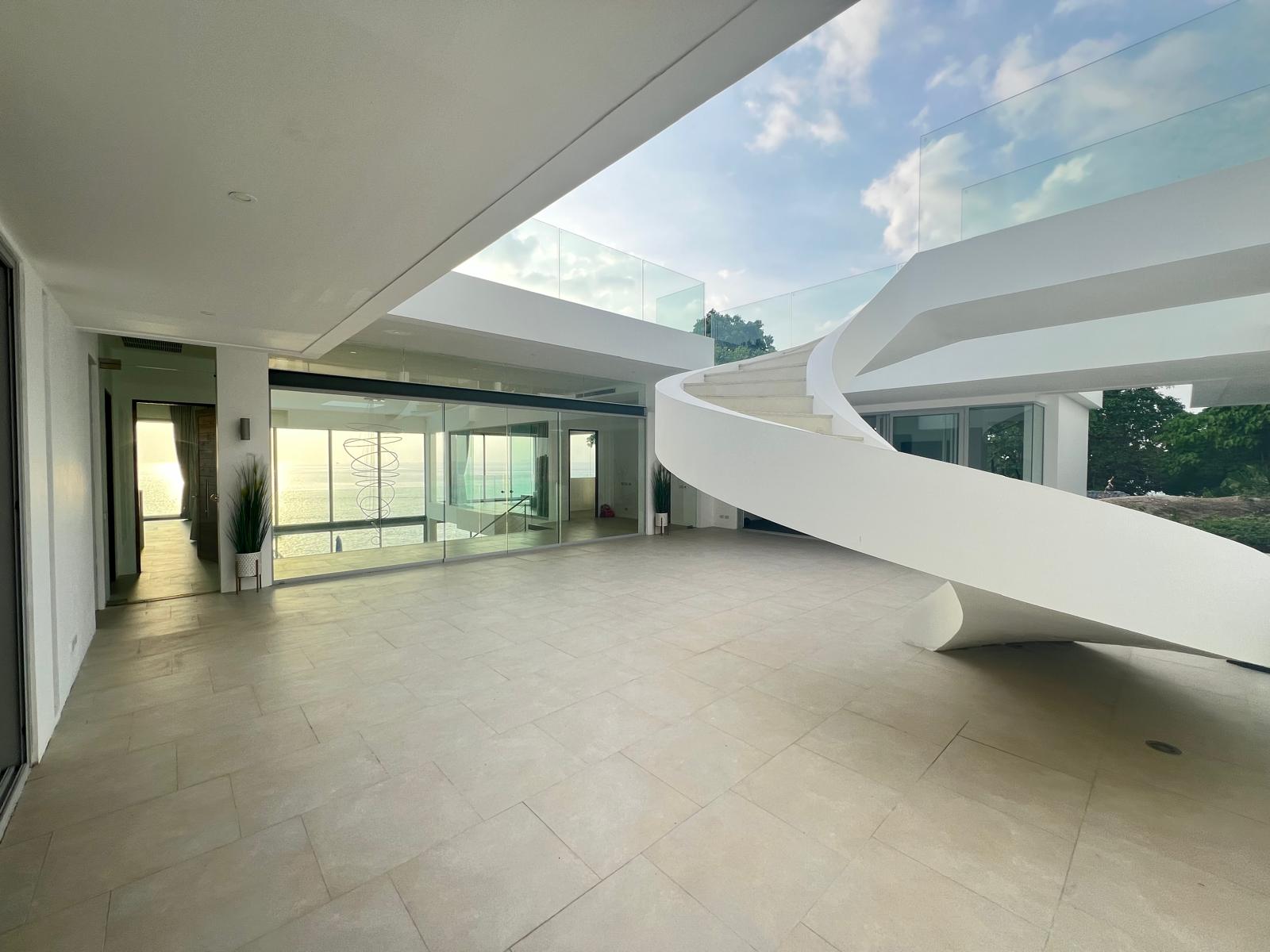 Open terrace of a modern white house with a spiraling white staircase, glass railings, and an ocean view above the horizon.