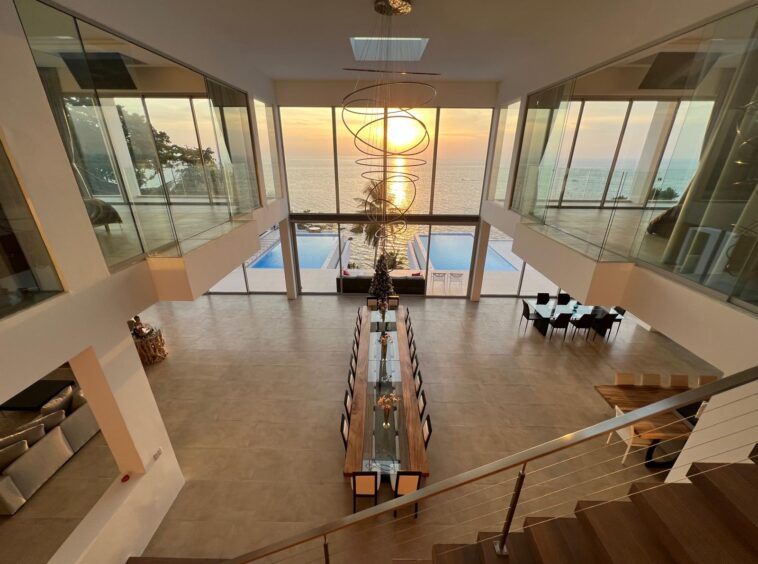 Modern two-story living and dining area with a long glass-top table, overlooking a sunset ocean view through large glass walls.