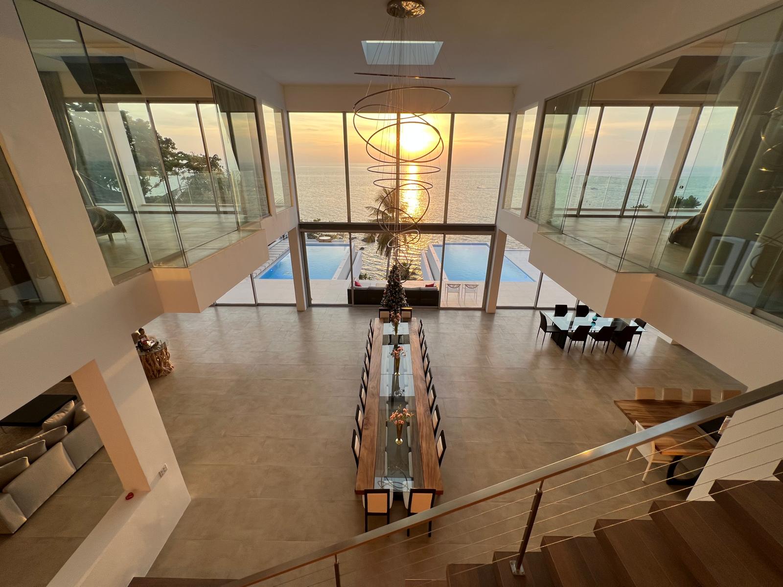 Modern two-story living and dining area with a long glass-top table, overlooking a sunset ocean view through large glass walls.