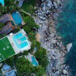 Aerial view of a seaside villa cluster with turquoise pools, lush greenery, and a rocky shoreline meeting blue ocean.