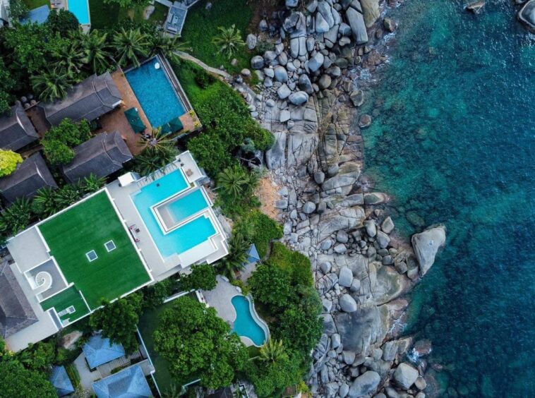 Aerial view of a seaside villa cluster with turquoise pools, lush greenery, and a rocky shoreline meeting blue ocean.