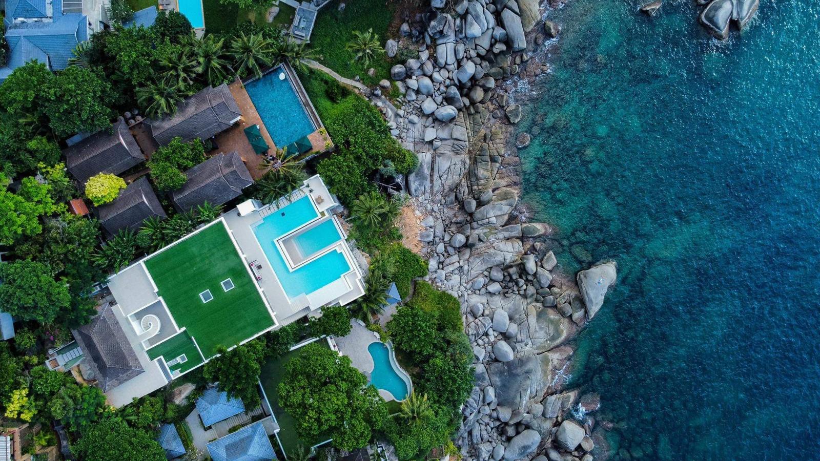 Aerial view of a seaside villa cluster with turquoise pools, lush greenery, and a rocky shoreline meeting blue ocean.