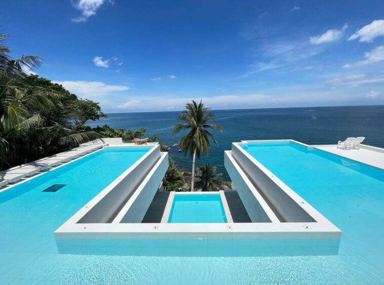 Modern white terrace with two turquoise infinity pools overlooking the blue ocean and palm trees under a clear sky.