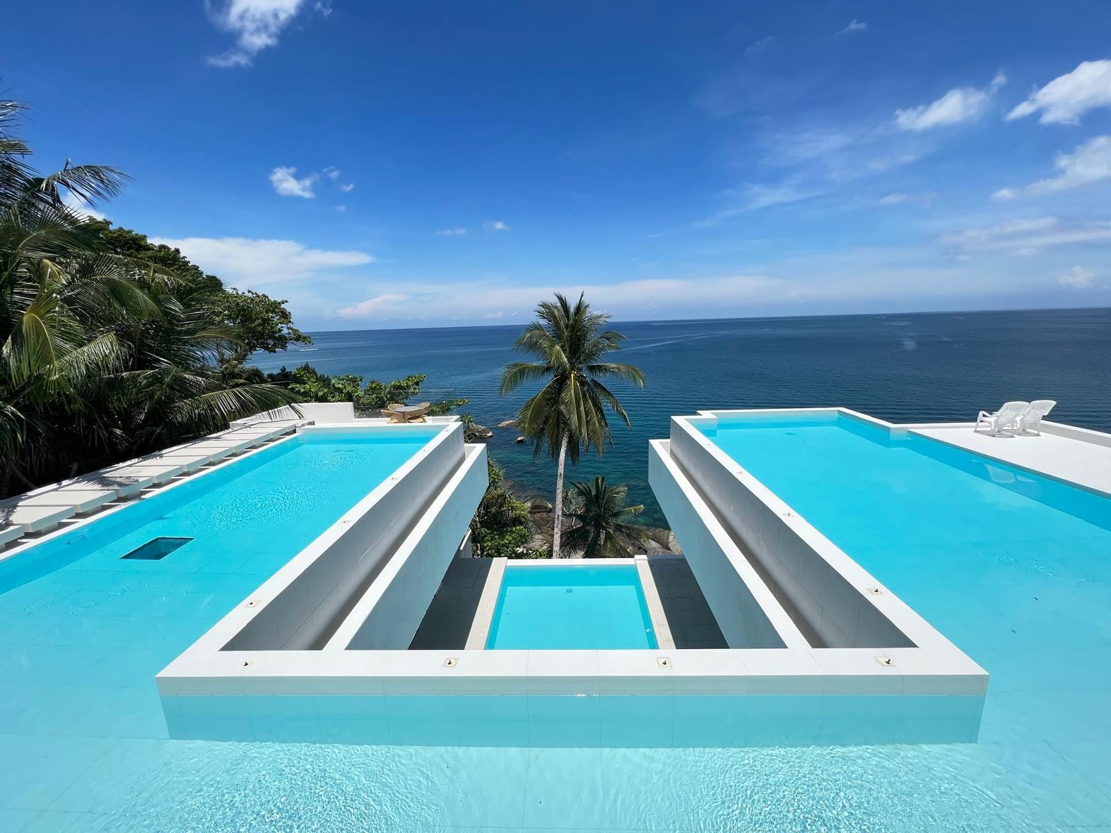 Modern white terrace with two turquoise infinity pools overlooking the blue ocean and palm trees under a clear sky.