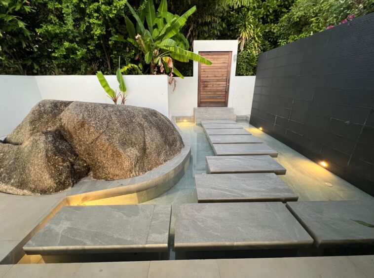 Modern outdoor courtyard with a stepping-stone path over a shallow pool toward a wooden door, large rock sculpture on the left, and lush greenery.
