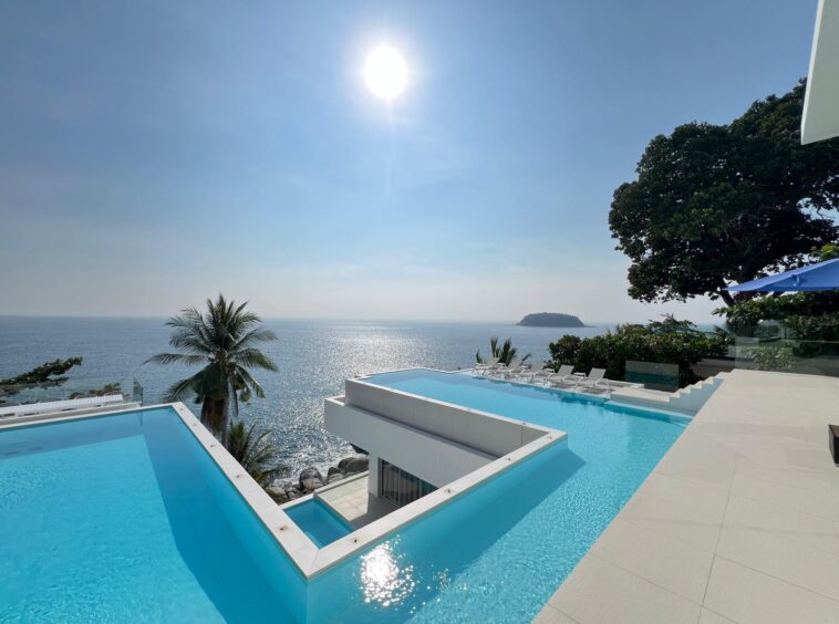Infinity pool terraces overlooking the sea with palm trees, lounge chairs, and a distant island on the horizon.