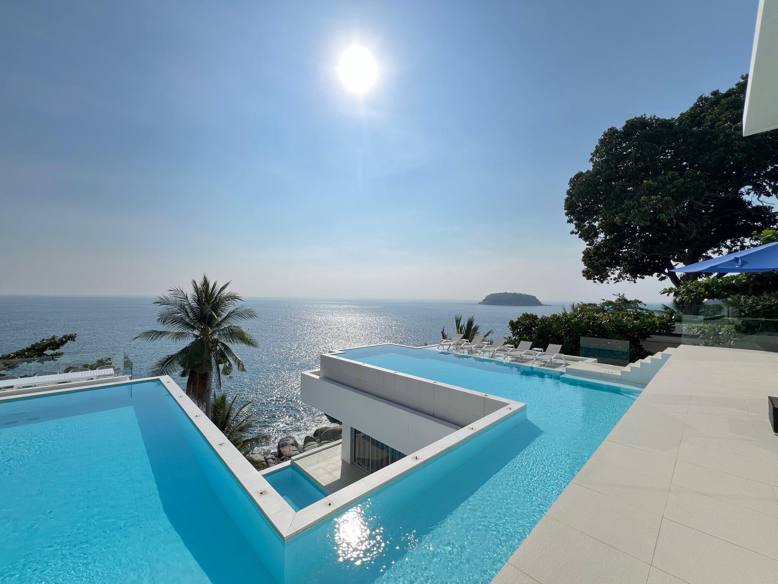 Infinity pool terraces overlooking the sea with palm trees, lounge chairs, and a distant island on the horizon.