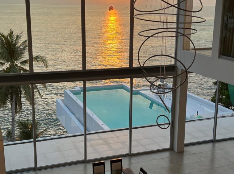 Open dining area with a long wooden table and glass centerpiece, large windows show a sunset over the sea and a rectangular pool outside.