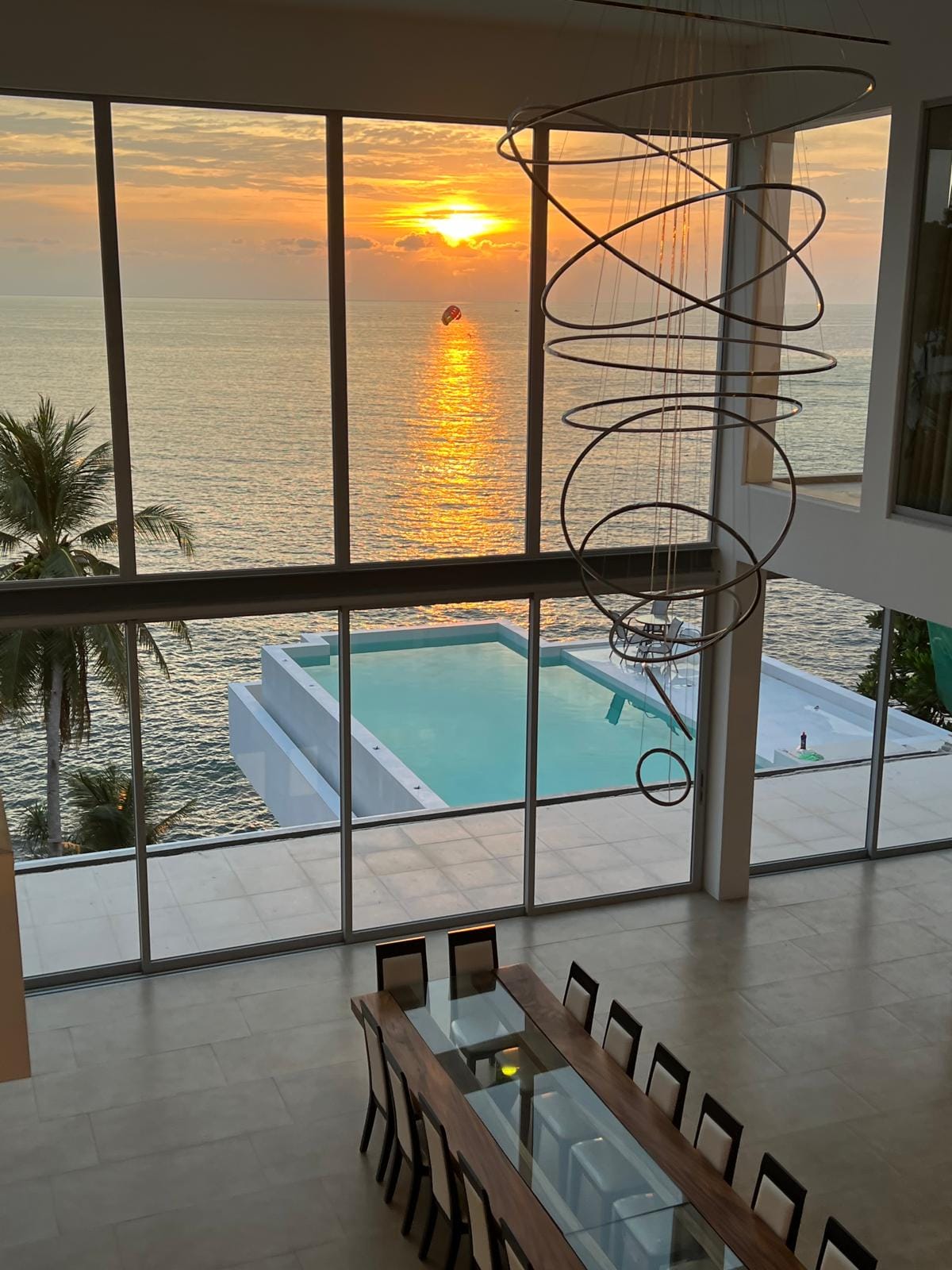 Open dining area with a long wooden table and glass centerpiece, large windows show a sunset over the sea and a rectangular pool outside.