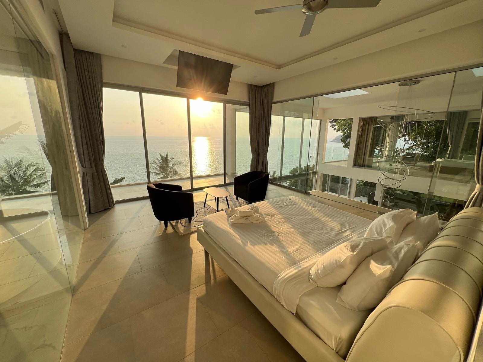 Sunset over the sea seen from a bright hotel suite with floor-to-ceiling windows, a white bed, and a seating area.