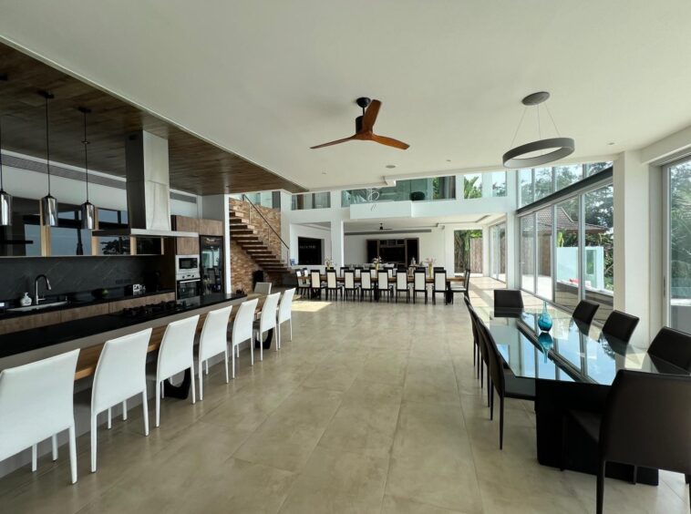 Open-concept kitchen and dining room with a long island, white bar stools, and glass-walled dining area bathed in natural light.