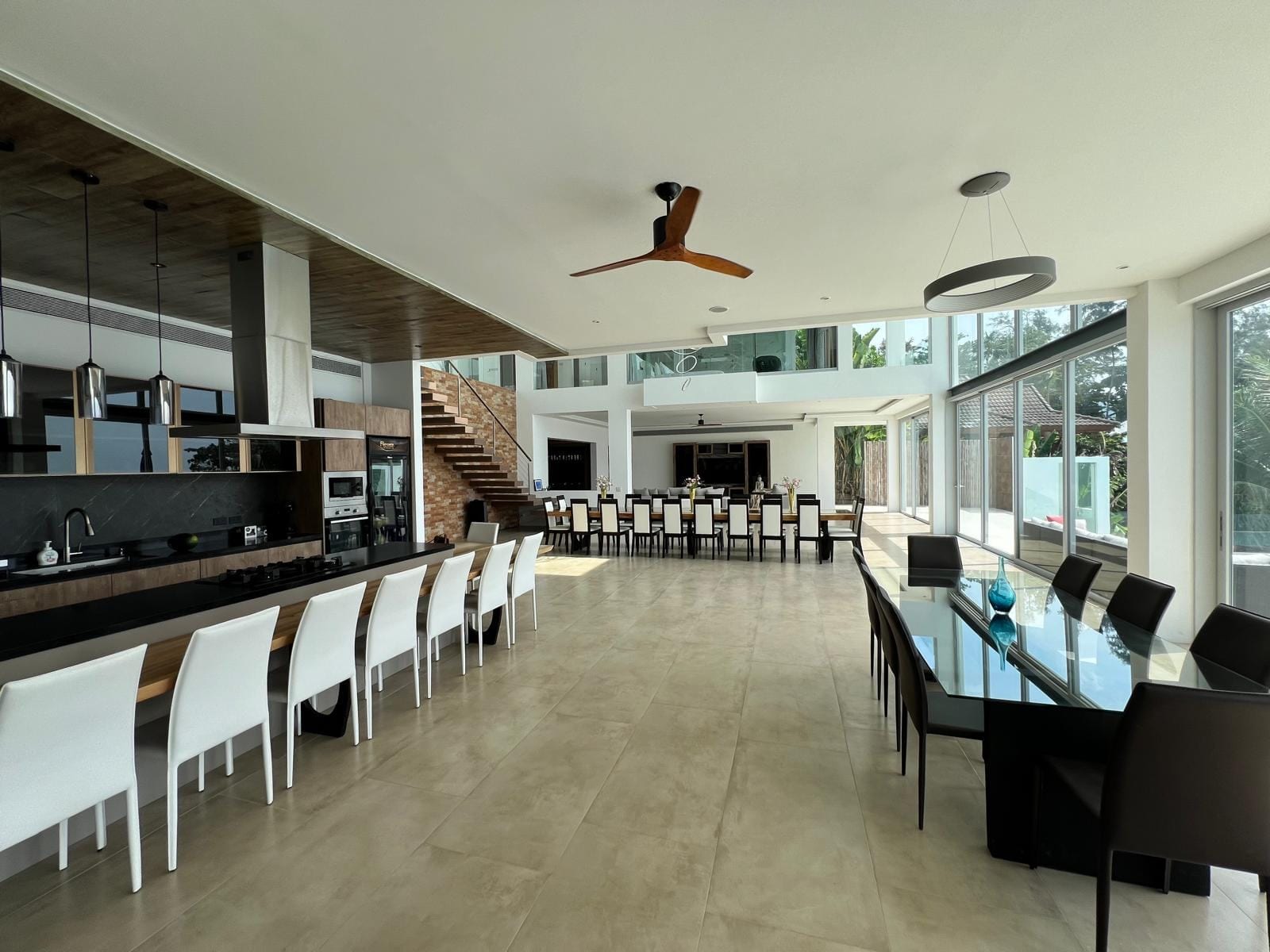 Open-concept kitchen and dining room with a long island, white bar stools, and glass-walled dining area bathed in natural light.