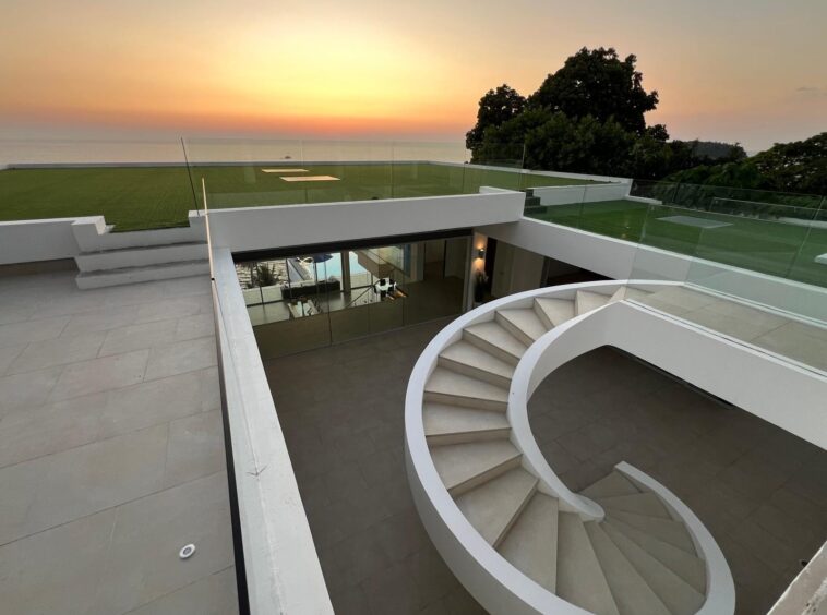 Modern terrace with a white spiral staircase overlooking a glass railing, sea sunset in the distance.