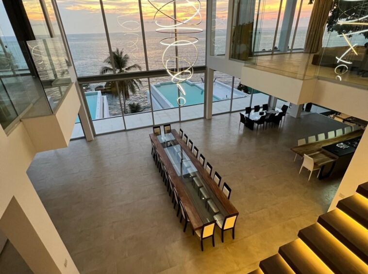 Two-story, modern dining area with a long wooden table and beige chairs, floor-to-ceiling windows overlook the sunset ocean, with a spiraling pendant chandelier and illuminated stairs nearby.