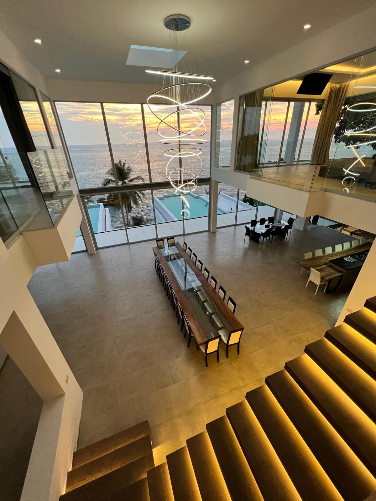Two-story, modern dining area with a long wooden table and beige chairs, floor-to-ceiling windows overlook the sunset ocean, with a spiraling pendant chandelier and illuminated stairs nearby.