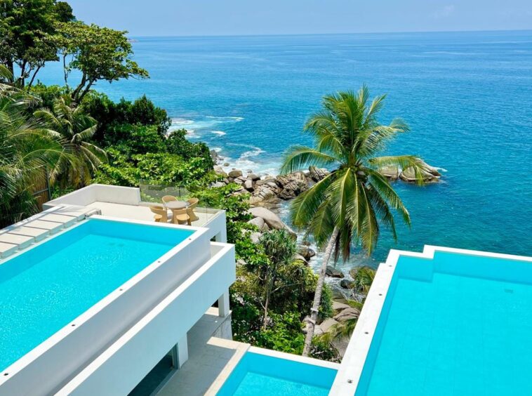 Aerial view of white, modern resort terraces with turquoise pools, overlooking the blue ocean and palm trees.