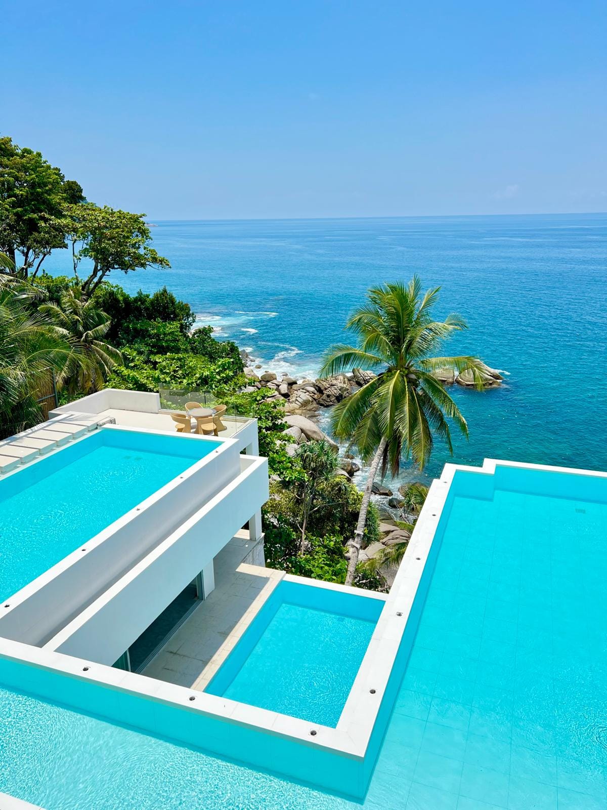Aerial view of white, modern resort terraces with turquoise pools, overlooking the blue ocean and palm trees.