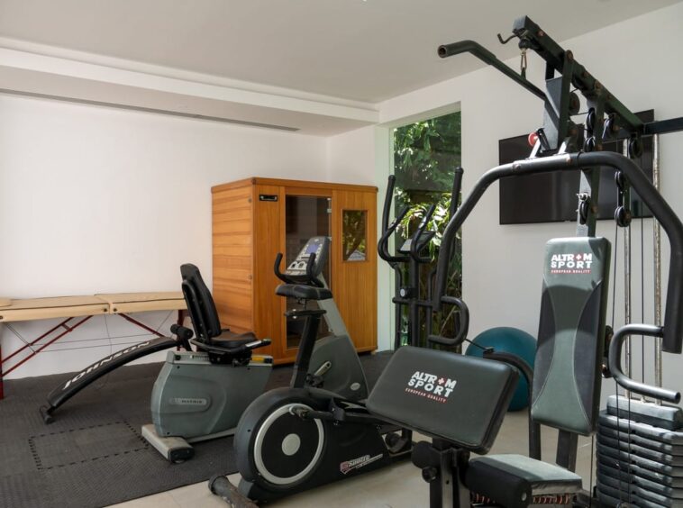Home gym with a multi‑station weight machine, an exercise bike, and a treadmill-like machine on padded flooring; a wooden cabinet against the wall.