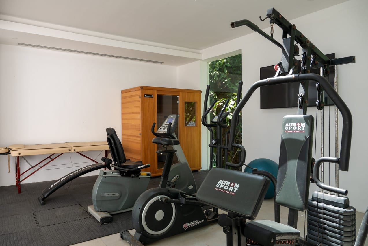 Home gym with a multi‑station weight machine, an exercise bike, and a treadmill-like machine on padded flooring; a wooden cabinet against the wall.